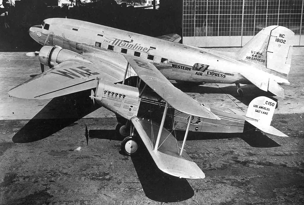The pictures has two aircraft parked in front of a hanger. In front is a Douglas M-2 biplane that is dwarfed by the Douglas DC-3 behind it. Both planes are painted in Western Air Express color schemes, the M-2 is painted red and silver with white lettering, the DC-3 silver with red lettering.