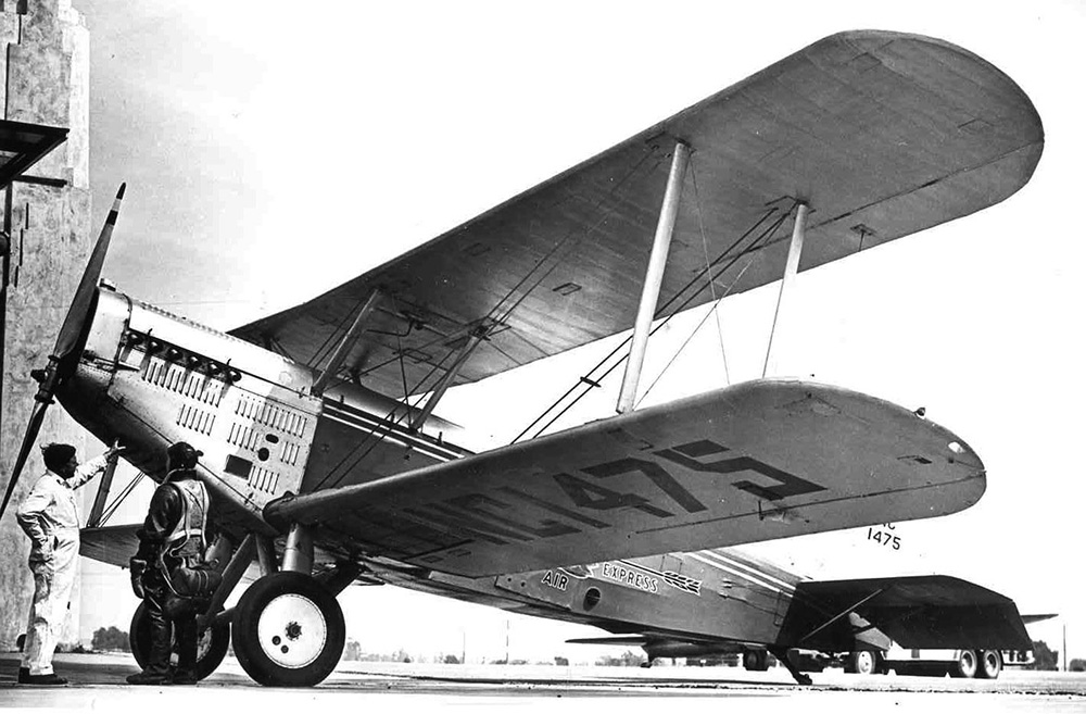 A Douglas M-4 biplane is center in picture. The plane sits at an airfield. The mechanic and pilot stand under the nose of the plane in conversation. The plane has the Wester Air Express Company logo on the side.