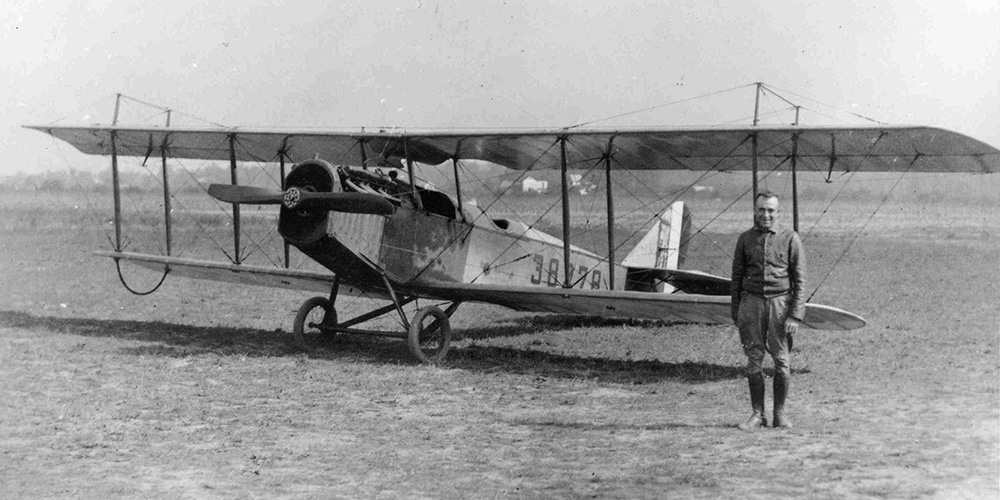 Air Mail Service pilot Eddie Gardner poses with a JN-4H biplane. The plane is parked in a field with Gardner standing with his arms at his sides smiling for the camera. The number 38278 is painted on its side.