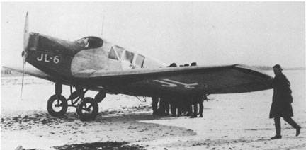 A monoplane marked JL-6 is parked in an icy field. Five men huddle at the back of the plane while one man walks to the front. The plane is an all metal plane.