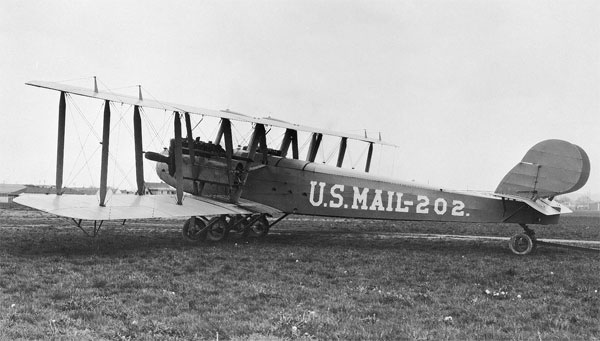 A Martin MB-1 in a field. The plane has the words U.S. Mail 202 on the side of the plane. The plane is very large and requires six wheels to support it, four under the wings and two under the tail.