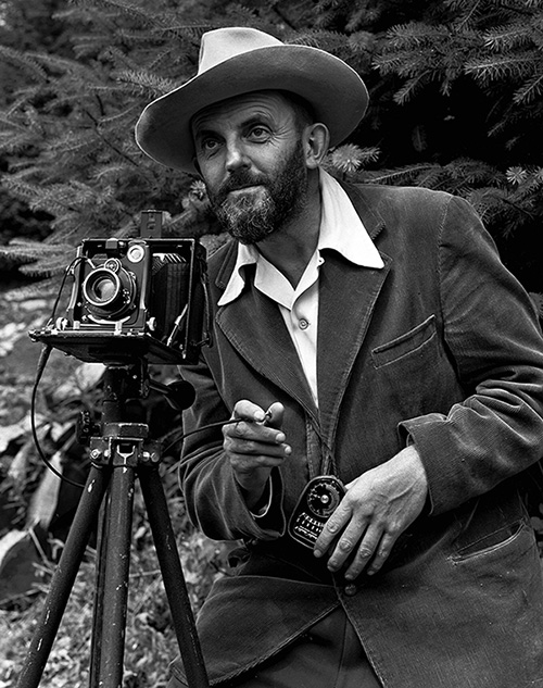 A photo of a bearded Ansel Adams with a camera on a tripod and a light meter in his hand. Adams is wearing a dark jacket and a white shirt, and the open shirt collar is spread over the lapel of his jacket. He is holding a cable release for the camera, and there is a rocky hillside behind him.