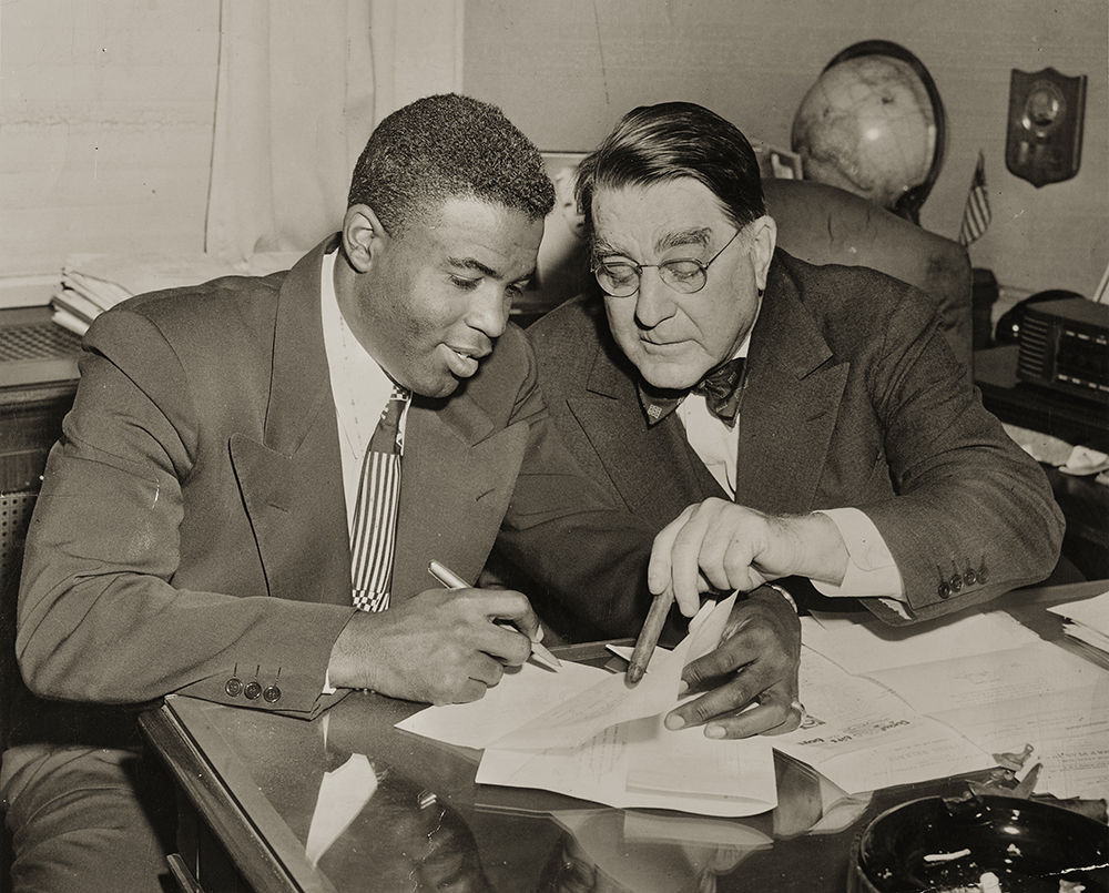 Jackie Robinson and Branch Rickey sitting at a desk looking at a contract.