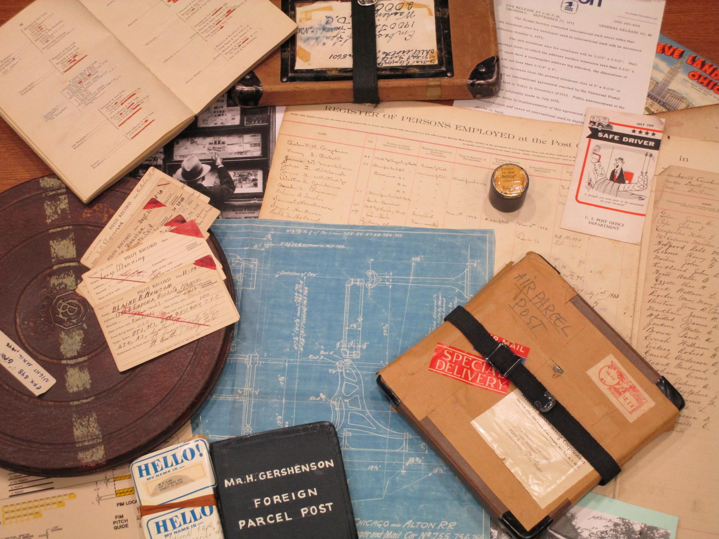 Photograph of various archival items such as registry of postal employees, nametags, brown paper parcel, notebooks, scrapbooks, photographs and other documents scattered across a desk