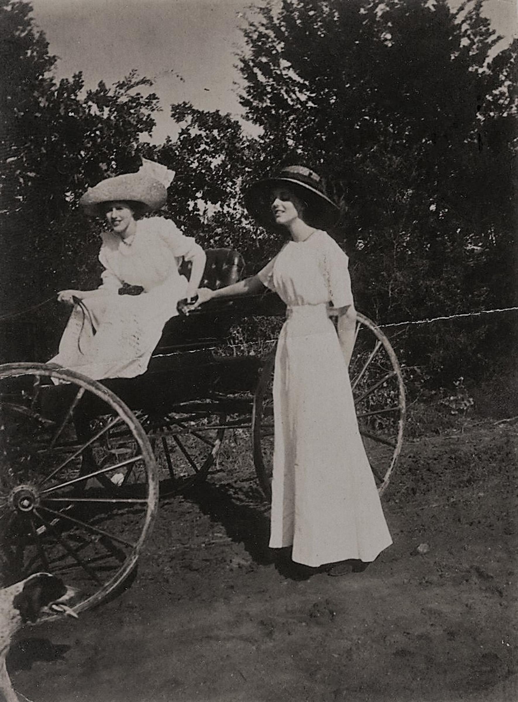 Two women in long old fashioned dresses and hats with a carriage.