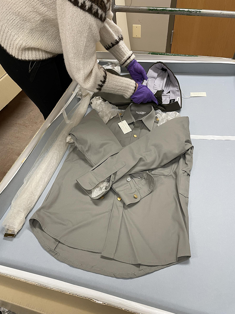 Assistant Curator Alison Bazylinski surveys a postal uniform. An individual bent over a flat surface with a blue tray that contains a pair of dark trousers, gray shirt, and belt wrapped in tissue. The individual is folding over the gray trousers.