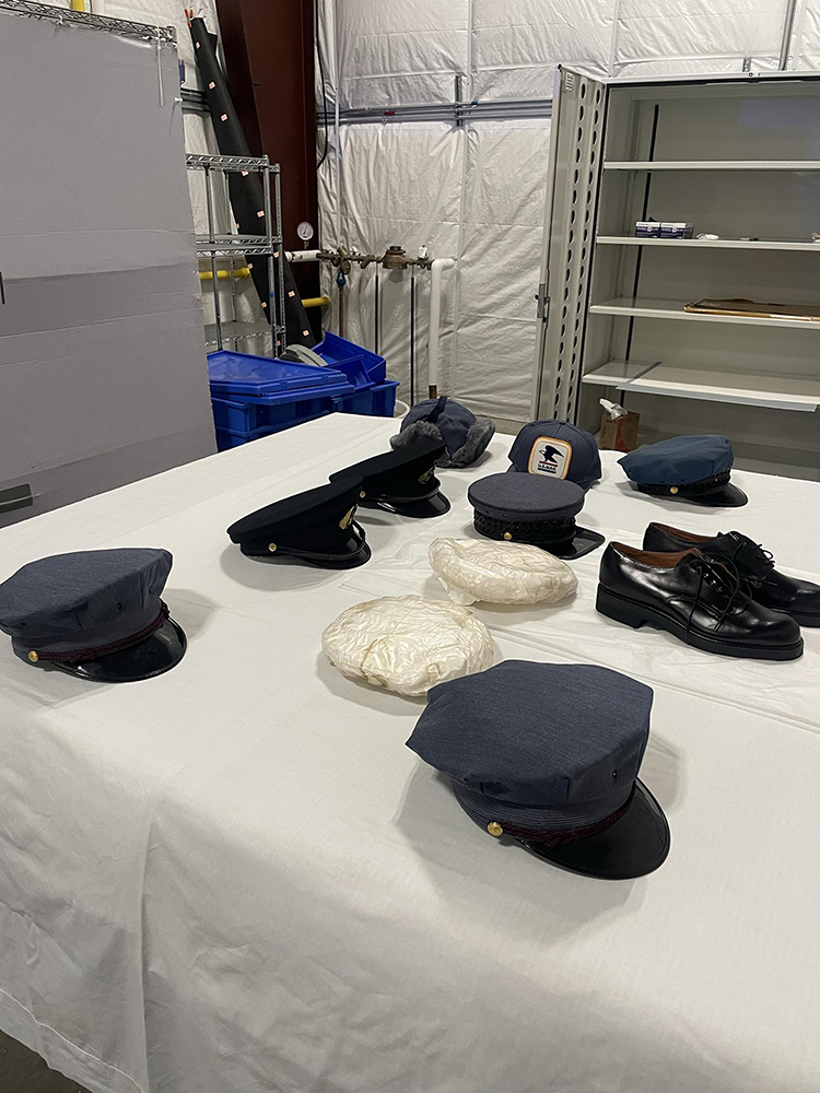 The contents of one box pulled for review and cataloging. A room with an open steel cabinet and a table covered with a sheet. 8 hats, two plastic caps, and a pair of shoes sit on the sheet-covered table.