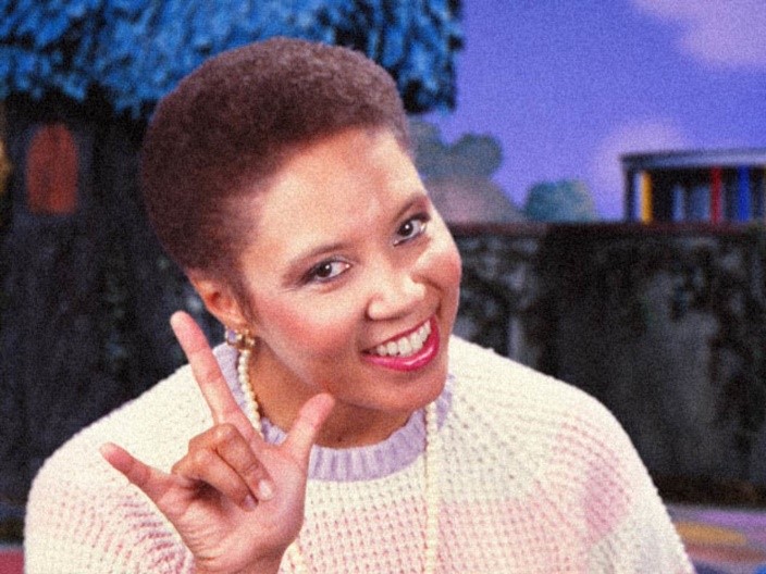 Photograph of a woman (Maggie Stewart) smiling and making the ASL sign for “I love you” with her right hand. 