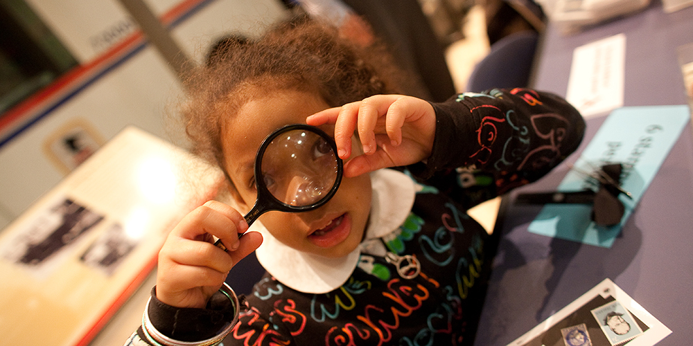 a child in a colorful shirt looks through a magnifying glass at the viewer.