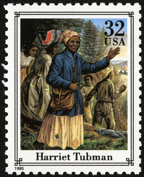 a 32-cent stamp with Harriet Tubman, an African American woman, in the foreground. She is wearing a blue coat and carrying a satchel. She has her left hand up, gesturing to four people behind her, who are looking through trees.