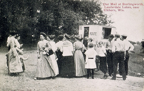People on a dirt road happily surround the rural mail carrier to receive their items.