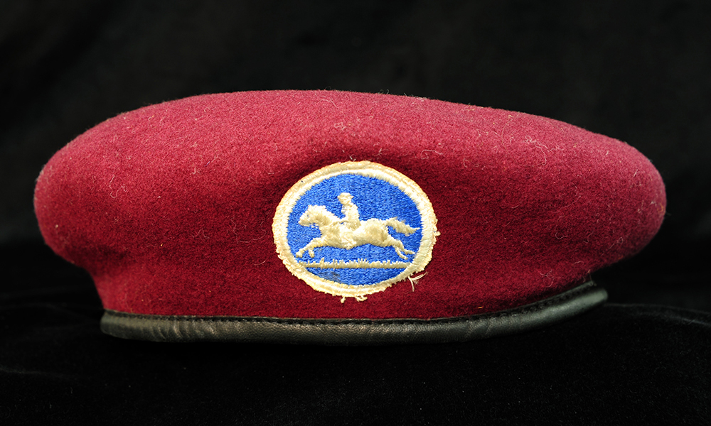 a wool, bright red beret style hat with black leather trim along the edge. In the center is a patch with a blue background, a horse and rider embroidered in white, with a white ring around the edge.