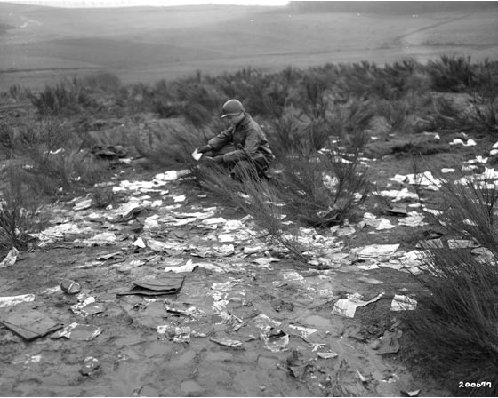 Tank soldier stoops looking at a letter in a field strewn with the contents of a mail sack.