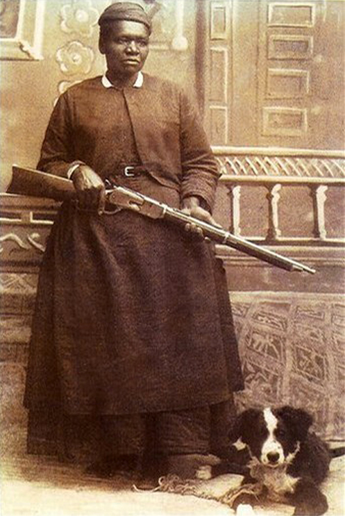 Black and white photograph of standing woman holding rifle with dog at her feet